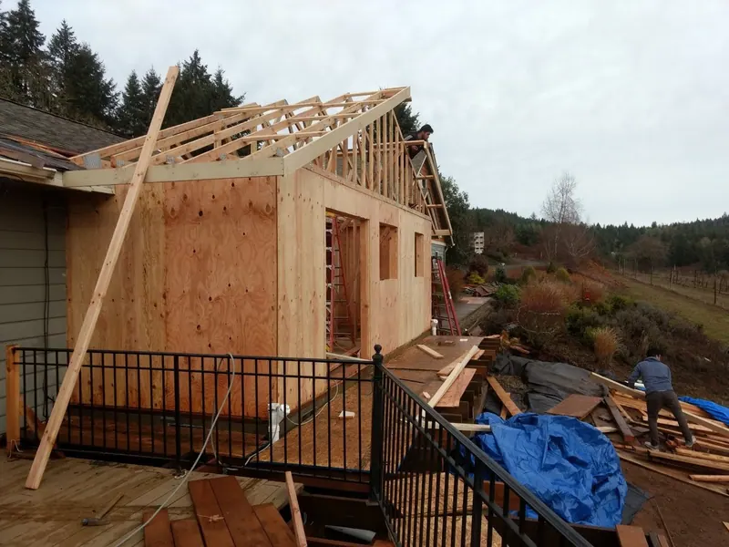 Workers set roof trusses on the OSB-sheathed walls of a new home addition on a cloudy Oregon day.