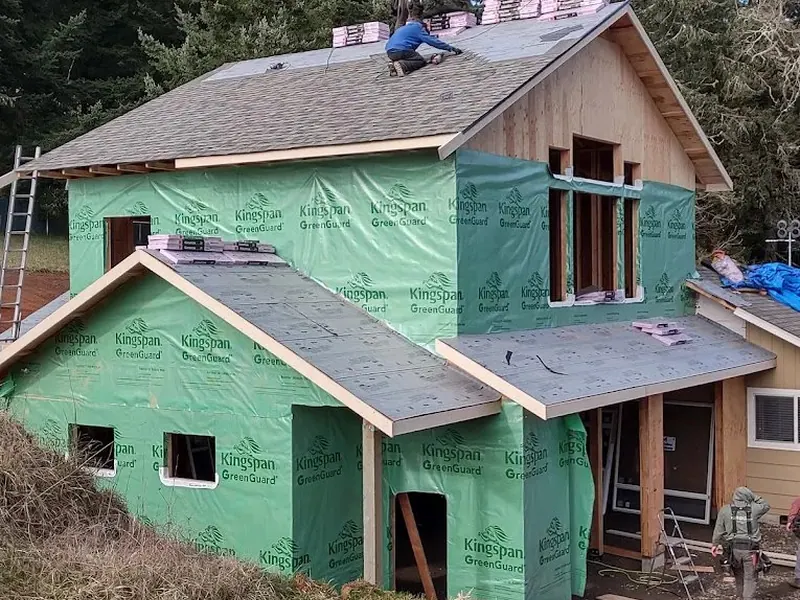 Two-story home addition wrapped in Kingspan GreenGuard with roofers installing asphalt shingles, a Red Umbrella home addition project in the Pacific Northwest.